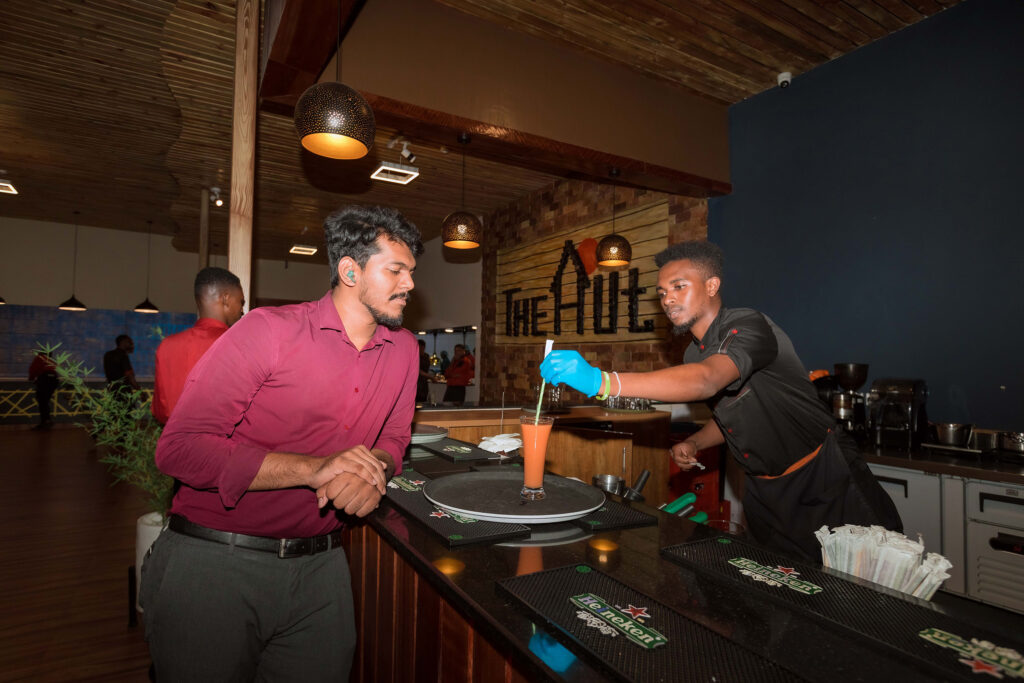 A bartender at The Hut in Kigali prepares a cocktail for a customer at the bar, creating a vibrant nightlife atmosphere with warm lighting and stylish decor